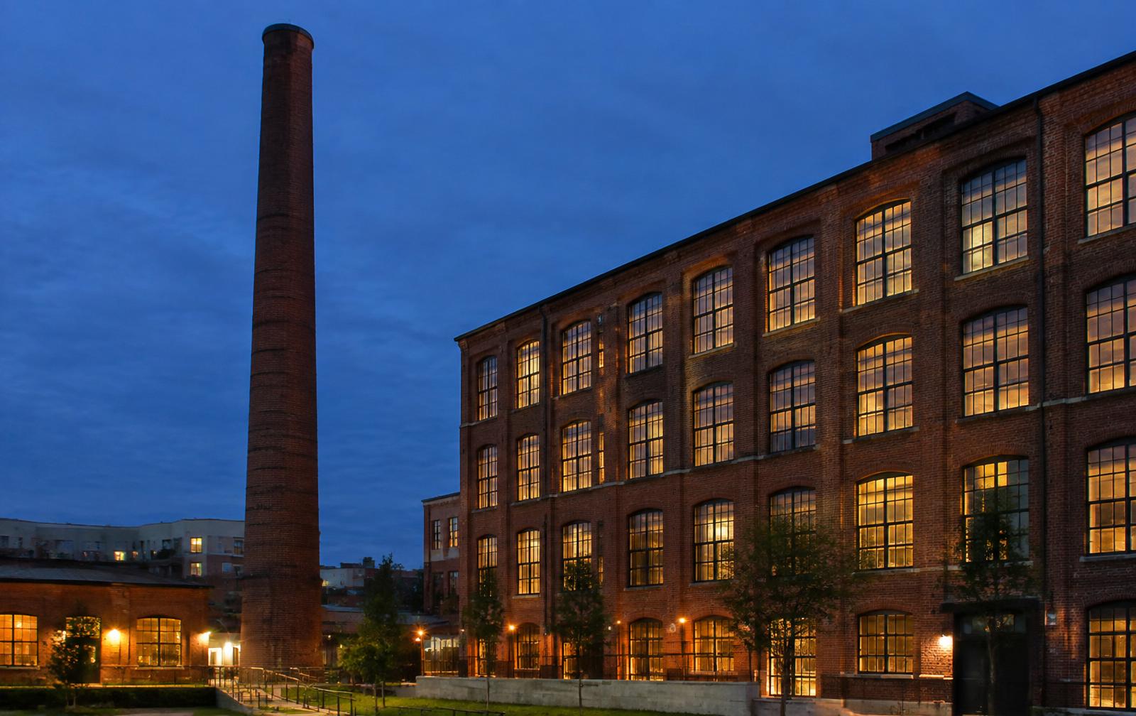 A brick mill smokestack and lit loft windows at blue hour in Belmont's Chronicle District